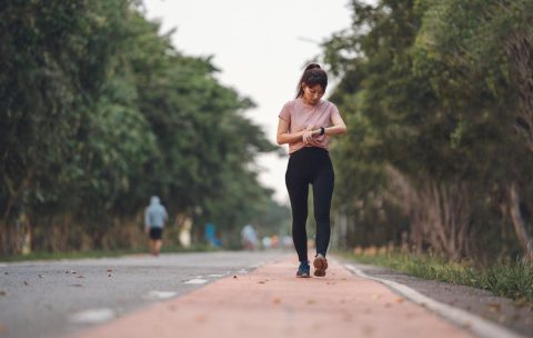 Young fitness woman runner checking time from smart watch. Young woman checking heart rate while jogging on street at dusk. Young woman looking on smartwatch her heartbeat while running in city.